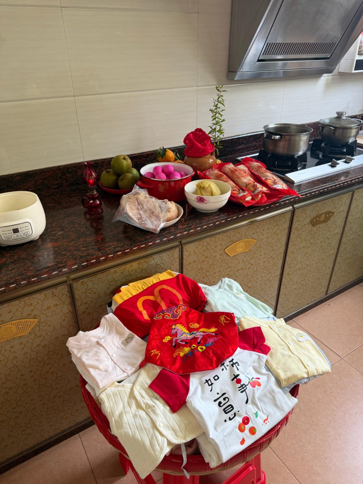 满月摆出的东西:成摞红团、一只鸡、橘与红蛋,旁边椅子上摊着婴儿新衣和绣花红肚兜。图源:抖音 @用户1284995464545忘忧草,7 月 12 日。